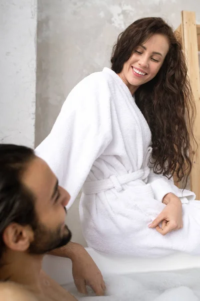 穿着白色浴衣的快乐的年轻女人坐在丈夫面前的浴缸里和他说话 — 图库照片