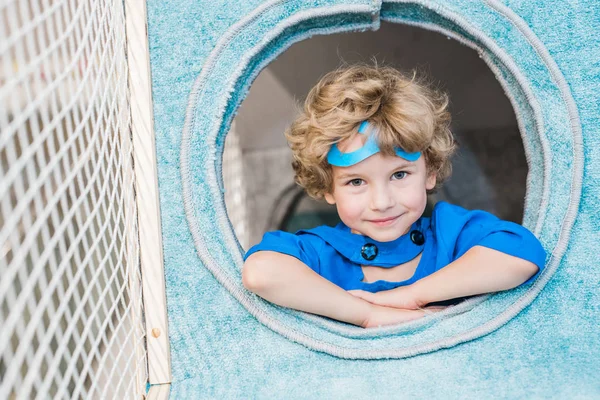 金发卷曲的小男孩穿着超人的蓝色服装 一边看着你 一边在运动场上的软房子里玩耍 — 图库照片