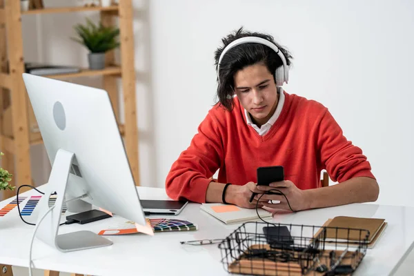 自宅でコンピュータ画面の前で仕事中にヘッドフォンで音楽を聞いている間にスマートフォンでスクロール深刻な男 — ストック写真
