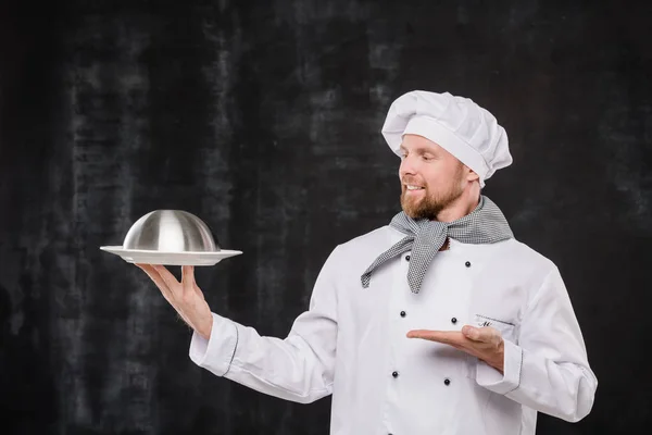 Young Smiling Chef Uniform Looking Pointing Cloche Cooked Meal While — 스톡 사진