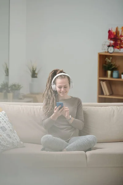 Chica Feliz Sentada Sofá Desplazándose Teléfono Inteligente Mientras Escucha Música — Foto de Stock