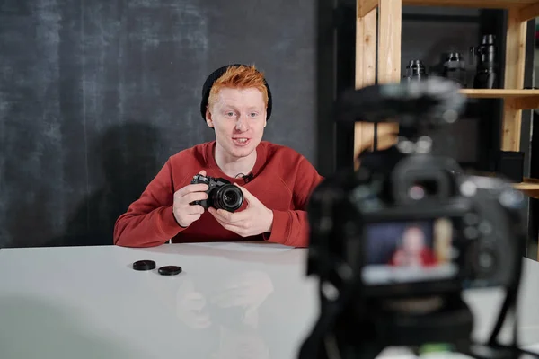 Joven Alegre Vlogger Por Escritorio Sosteniendo Una Nueva Cámara Fotográfica — Foto de Stock