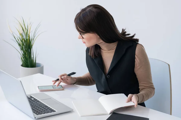 若い成功したビジネス女性とともにオープンコピーブックスクロールスマートフォンノートパソコンの前に座っている間 — ストック写真