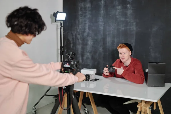 若いオペレーターは スタジオでオンライン視聴者にそれを示しながら 新しいフォトカメラを指して幸せな男性Vloggerを撮影します — ストック写真