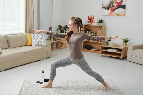 Fit girl in leggins and pullover stretching legs and arms while exercising on carpet during workout at home