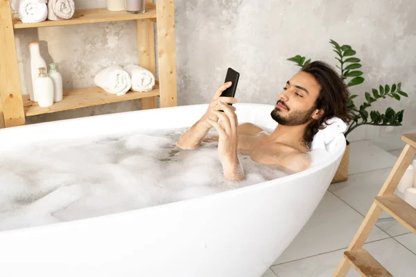 Jovem Homem Assistindo Vídeo Rolagem Smartphone Enquanto Deitado Banheira Cheia — Fotografia de Stock
