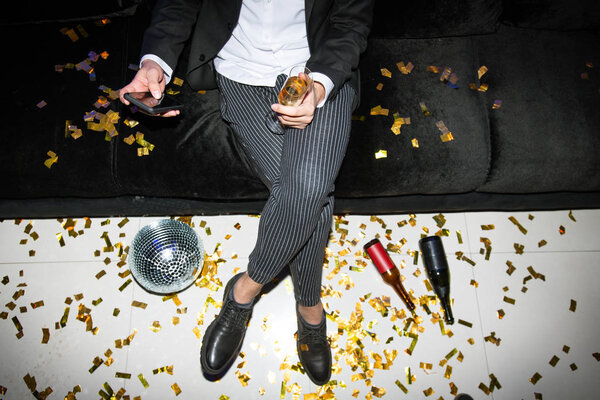 Low section of young elegant man with smartphone and flute of champagne relaxing on couch and texting