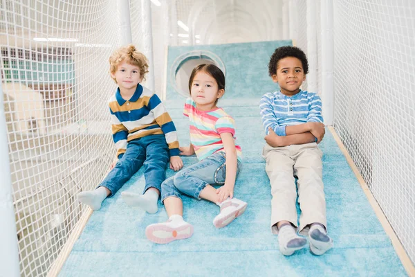 レジャーセンターの遊び場に座っている間 あなたを見ている3人の愛らしい異文化間の子供たちのグループ — ストック写真