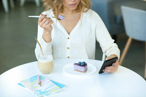 年轻而随意的女人在智能手机里打滚 坐在餐桌旁 吃着蓝莓芝士蛋糕 — 图库照片