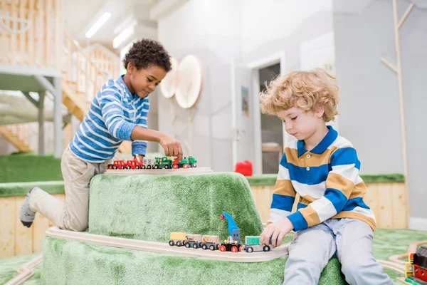 現代の子供センターで一緒におもちゃの列車をプレイしながら 遊び場に座っている2人の異文化間の男の子 — ストック写真