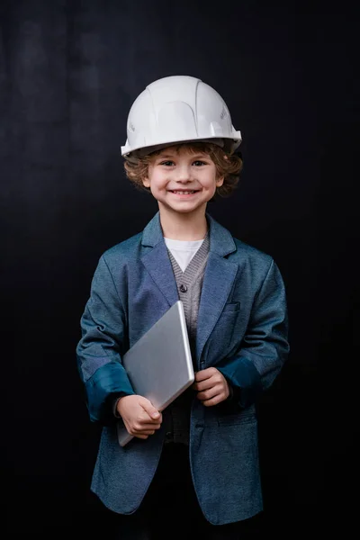 戴着硬礼帽 身穿正装的小男孩 拿着折叠的笔记本电脑 面带微笑地看着你 与黑色背景隔离在一起 — 图库照片