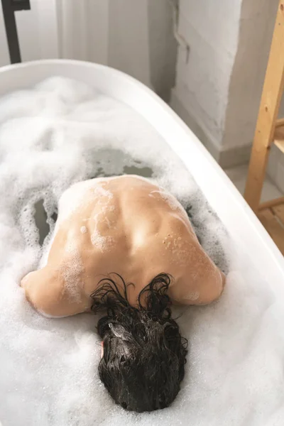 一个赤身裸体的年轻人坐在水里 一边用泡沫洗澡 一边洗身体 — 图库照片
