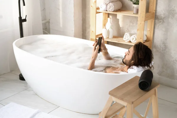 年轻而安详的男人躺在浴缸里 在浴室的角落里装满水和泡沫 一边用智能手机 — 图库照片