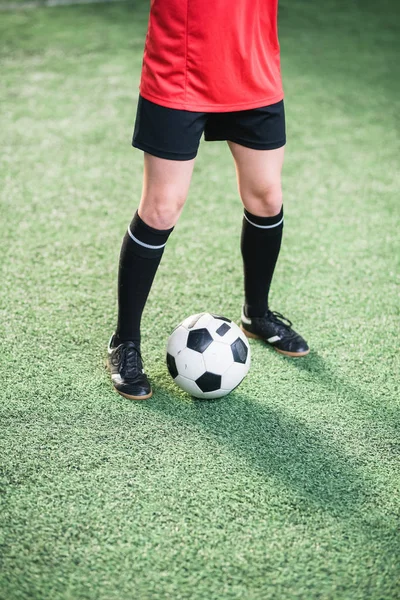 Soccer ball between legs of active girl in sports uniform standing on green football field during training