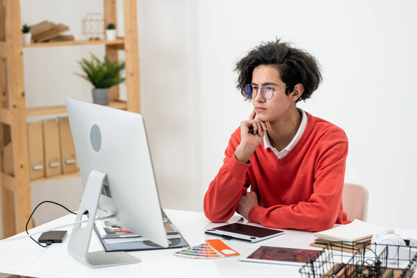 Serious web designer in eyeglasses looking through examples on computer screen while working over new logo by desk