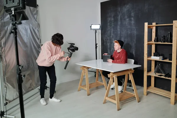 年轻的视频操作员穿着便服 拿着相机坐在博客作者面前 黑匣子坐在工作室的桌子旁 — 图库照片