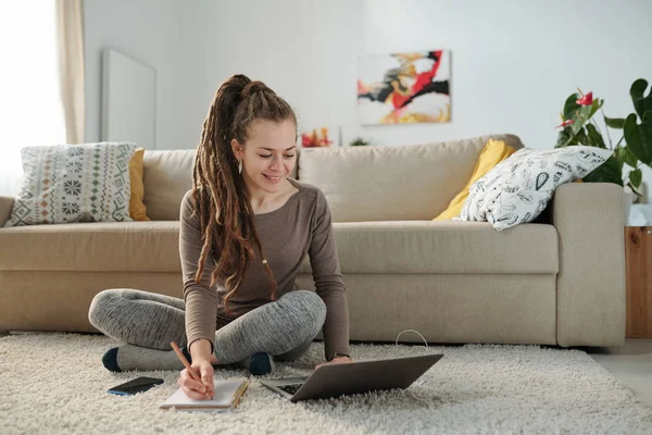 Cute Uśmiechnięta Studentka Dredami Robi Notatki Siedząc Podłodze Przed Laptopem — Zdjęcie stockowe