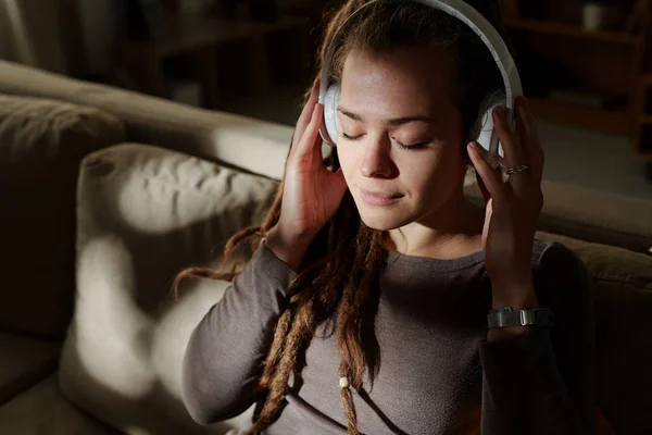 Calm young female with headphones enjoying music for sleep while sitting on couch in dark room