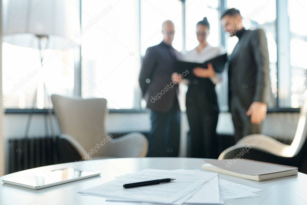 Touchpad, notebook, papers and pen on table on background of group of business partners having meeting