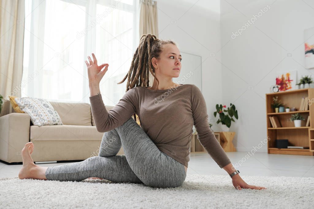 Young female with dreadlocks sitting on the floor in one of yoga positions while stretching arm and leg muscles