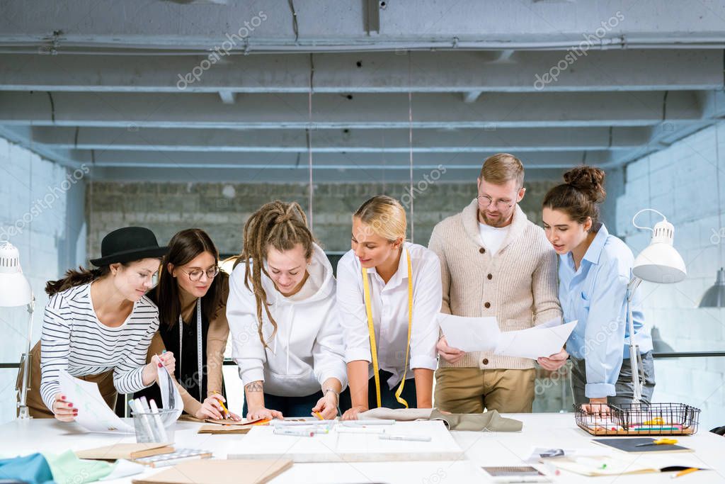 Group of six young creative designers bending over desk while discussing stuff for new fashion collection in studio