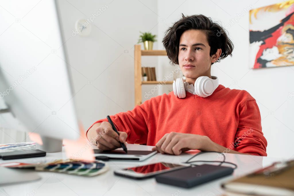 Young serious freelancer in casualwear working at home in front of computer screen and retouching photos