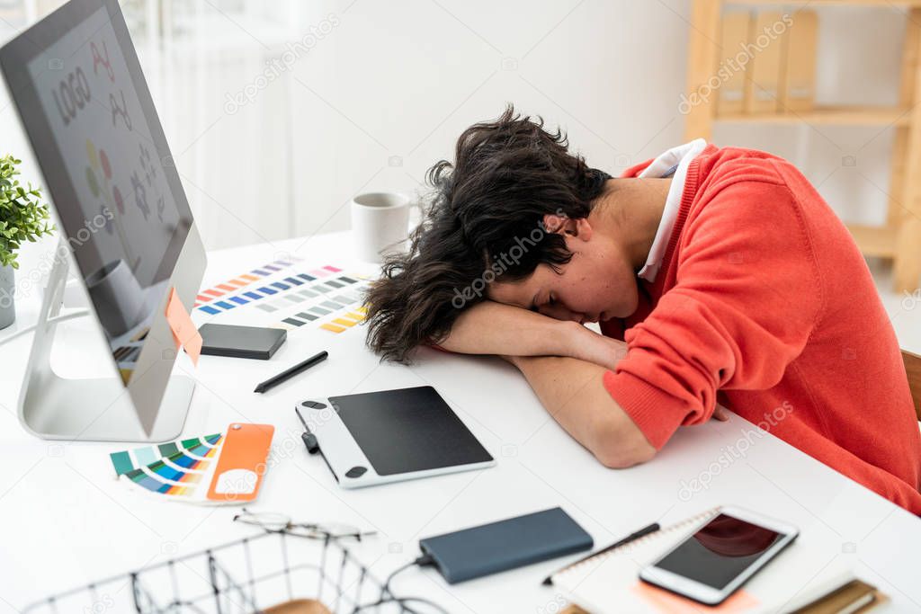 Exhausted freelancer napping on desk by computer among working supplies and gadgets after work