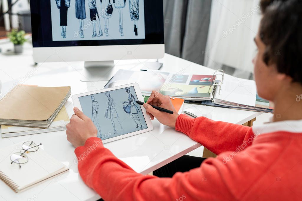 Young creative fashion designer with tablet scrolling through new models while sitting by desk in front of computer screen