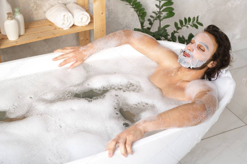 Serene young man with face mask relaxing in hot bath with foam while keeping his eyes closed