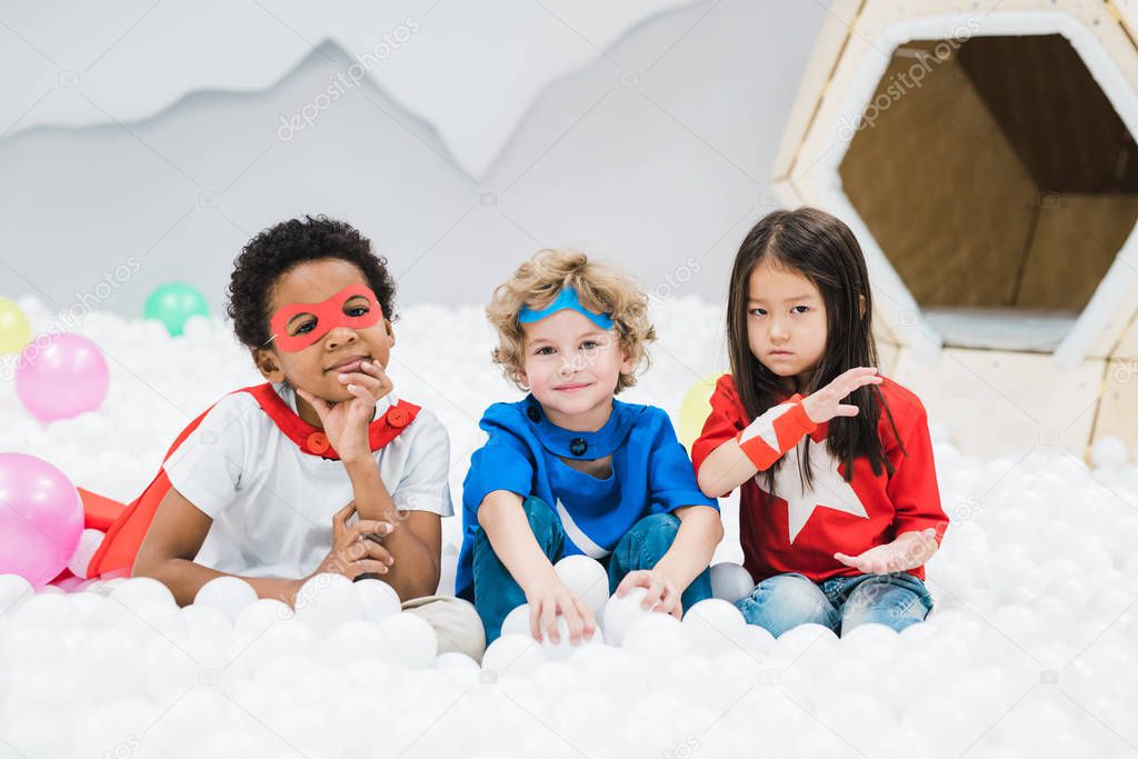 Group of adorable little intercultural friends in costumes sitting among white balloons in children room