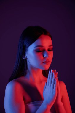 Sakin, çıplak omuzlu genç bir kadın. Gözleri kapalı meditasyon yapıyor. Koyu mavi arka plan, neon ışığı.