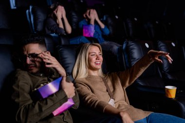 Genç, gülümseyen bir kadın, elinde patlamış mısır kutusuyla aksiyon filmi izlerken ekranı işaret ediyor.