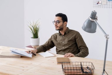 Masa başı ödevlerini hazırlarken gözlüklü genç bir öğrenci not defterindeki notlarına bakıyor.