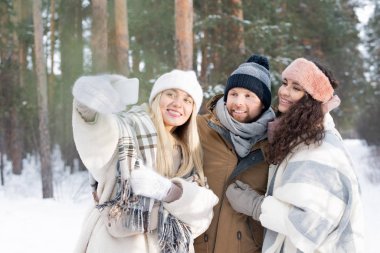 Üç neşeli ve sevecen arkadaş kış ormanlarında selfie çekerken gülümseyen akıllı telefon kameralarına bakıyorlar.