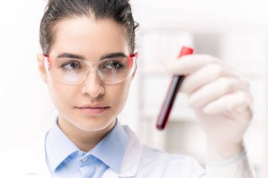 Laboratuvarda geliştirilen yeni aşı örneğiyle birlikte mataraya bakan koruyucu gözlüklü oldukça genç ve ciddi bir kadın.