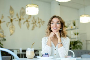 Gündelik kıyafetli, gülümseyen sarışın kadın kafenin yanında otururken bir bardak kapuçino ve cheesecake ile hayal kuruyor.
