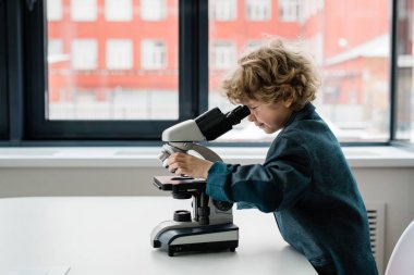 Laboratuar penceresiyle mikroskopta karşılaştırma yaparken günlük kıyafet giyen zeki bir öğrenci ya da biyolog.