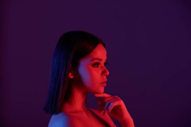 Neon ışıklarıyla çenesine dokunan düz saçlı, dalgın, çıplak genç bir kadın.