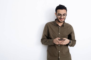 Gözlüklü ve gömlekli gülümseyen genç işadamı beyaz duvarın yanında duruyor ve akıllı telefonuyla mesajlaşıyor.