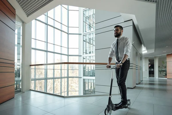 Happy young bearded businessman moving forwards while standing on scooter inside contemporary business center