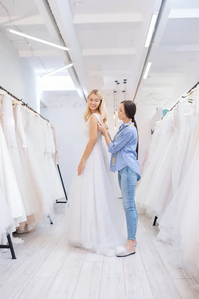 Smiling brunette wedding dress tailor assisting bride to wear dress between rows of beautiful gowns