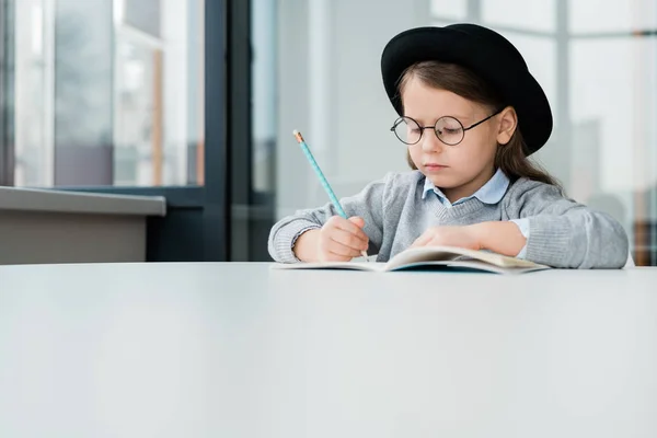 Ernsthaft Schlaues Schulmädchen Mit Hut Sitzt Schreibtisch Und Macht Notizen — Stockfoto