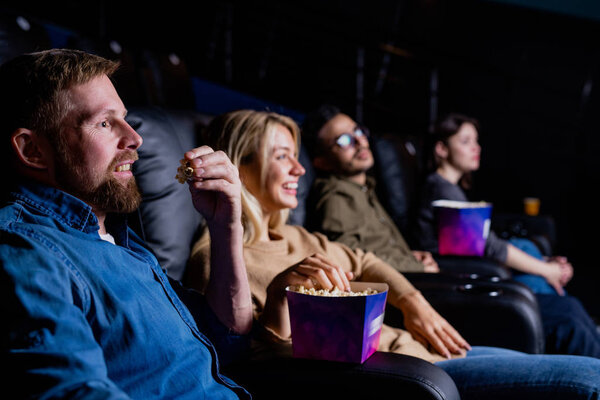 Row of happy young friends relaxing in comfortable armchairs in cinema while eating popcorn and watching movie