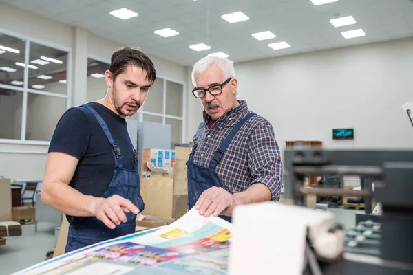 戴眼镜 触摸纸张 与同事谈论印刷厂印刷页的资深员工 — 图库照片