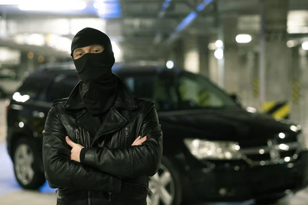 黒いジャケットの男性犯罪者と背景に駐車場に車で胸で腕を横断上のバラクラバ — ストック写真
