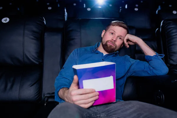 Young bored man with box of popcorn sitting on black leather armchair in front of large screen in cinema