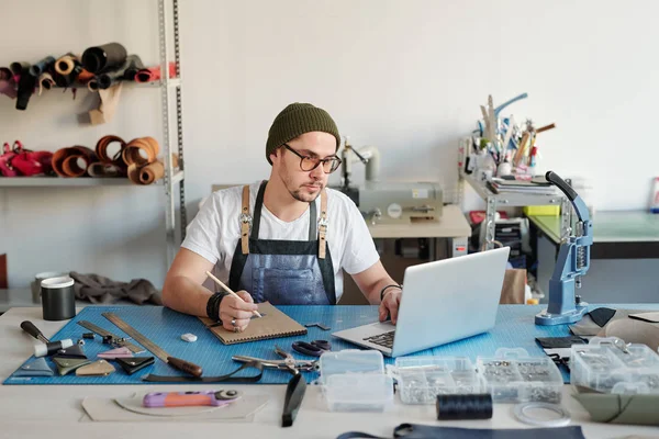 Jovem Sério Óculos Usando Laptop Fazendo Anotações Sketchpad Durante Processamento — Fotografia de Stock