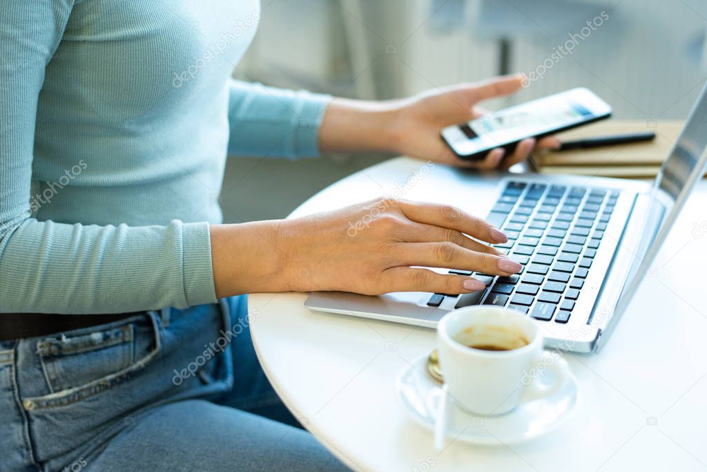 Hands of young contemporary freelancer or businesswoman touching keys of laptop keypad while using smartphone during work