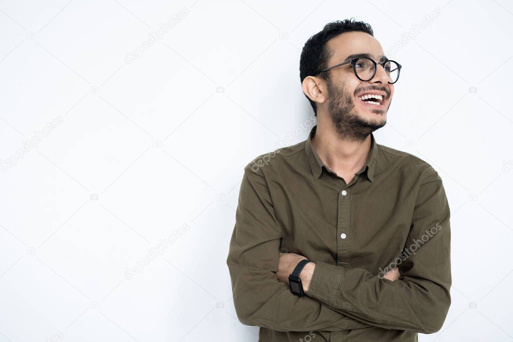Laughing guy in eyeglasses and casualwear crossing arms on chest while standing in isolation with copyspace on the left