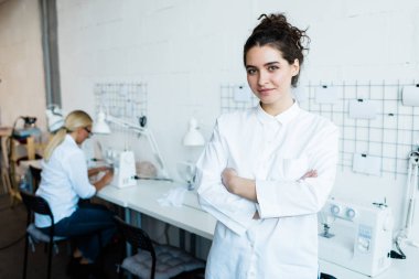 Uzun beyaz bluz giyen dikiş makinesi operatörünün portresi göğsünde iş yerine karşı kollarını kavuşturuyor